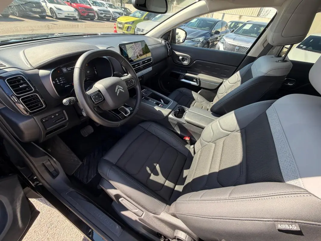 Vue intérieure côté conducteur du Citroën C5 Aircross Hybride 2024 noir, avec tableau de bord et sièges en tissu gris.