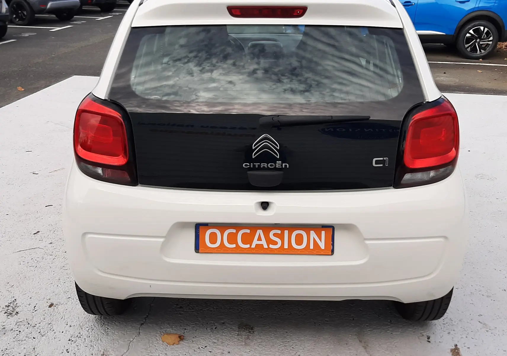 Vue arrière d'une Citroën C1 blanche 2021 avec hayon noir et plaque orange "OCCASION" sur un parking.