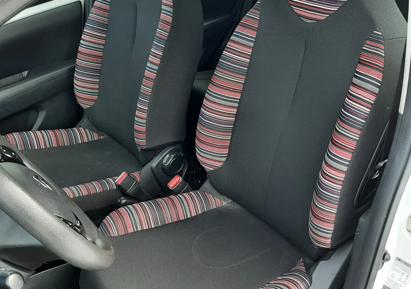 Vue intérieure côté gauche montrant les sièges avant en tissu noir avec motifs rayés rouges et gris d'une Citroën C1 blanche 2021.