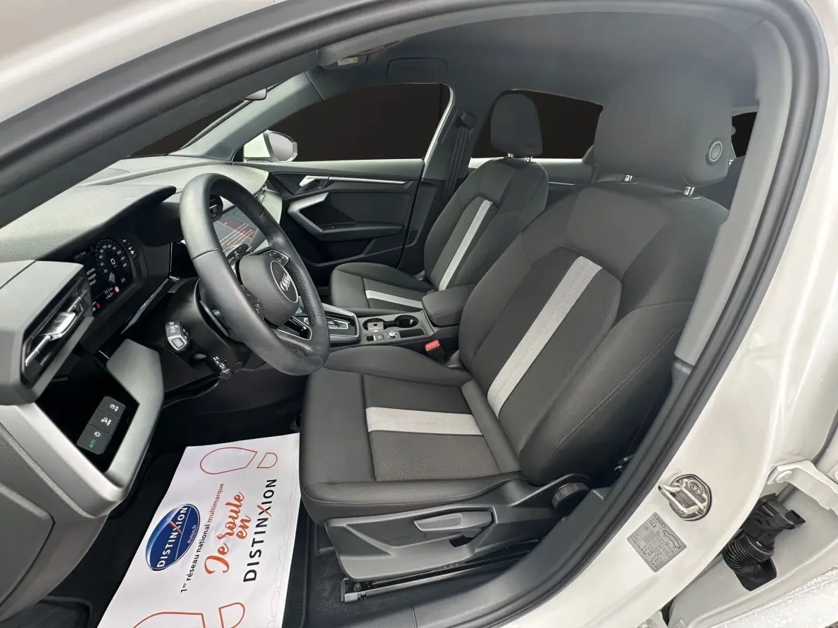 Vue intérieure côté conducteur de l'Audi A3 blanc 2024, sièges tissu noir avec bandes grises et tableau de bord moderne.