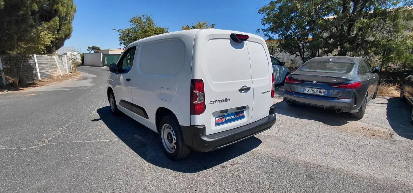 Vue 3/4 arrière droite d’un Citroën Berlingo blanc utilitaire avec porte latérale, garé sur une route ensoleillée.