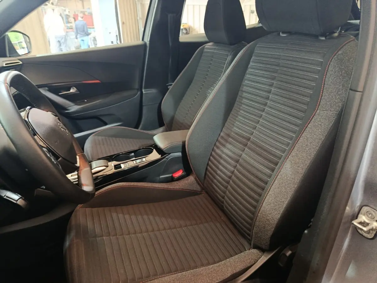 Vue intérieure côté conducteur du Peugeot E-2008 électrique gris clair, montrant les sièges avant en tissu avec surpiqûres rouges.