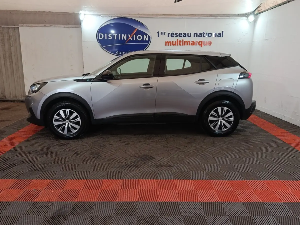 Profil côté gauche d'un Peugeot E-2008 électrique gris clair 2023, dans un showroom avec sol à damiers noir et rouge.
