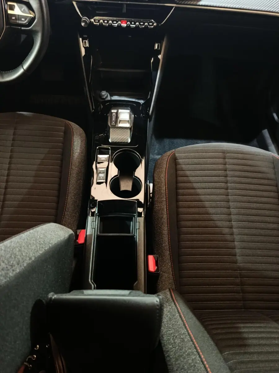 Vue intérieure centrée sur la console centrale noire brillante et les sièges tissu gris foncé avec surpiqûres rouges du Peugeot E-2008 électrique.