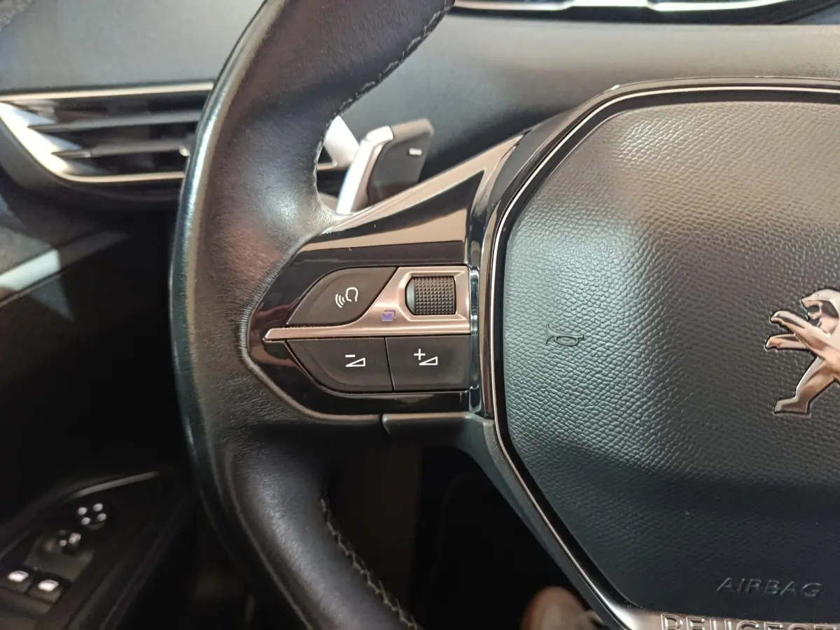 Gros plan sur le volant cuir noir du Peugeot 3008 avec commandes audio et logo lion visible au centre.