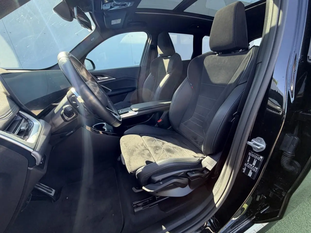 Vue intérieure côté conducteur du BMW X1 noir saphir, sièges sport en Alcantara avec surpiqûres bleues et toit panoramique visible.