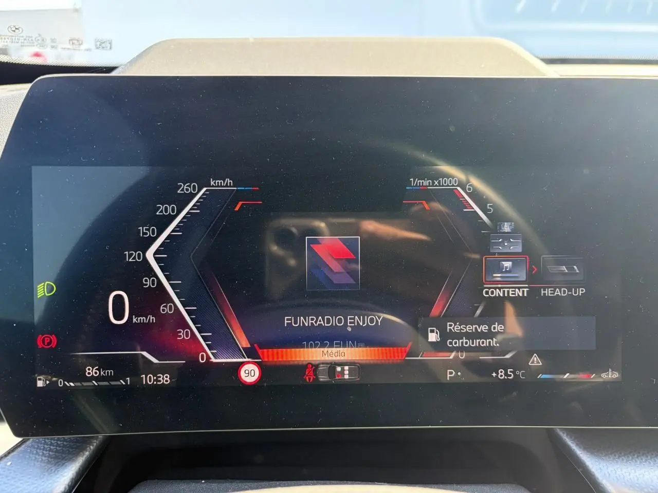 Gros plan sur le combiné d'instrumentation numérique du BMW X1 noir, affichant la vitesse, la radio et l'alerte réserve de carburant.