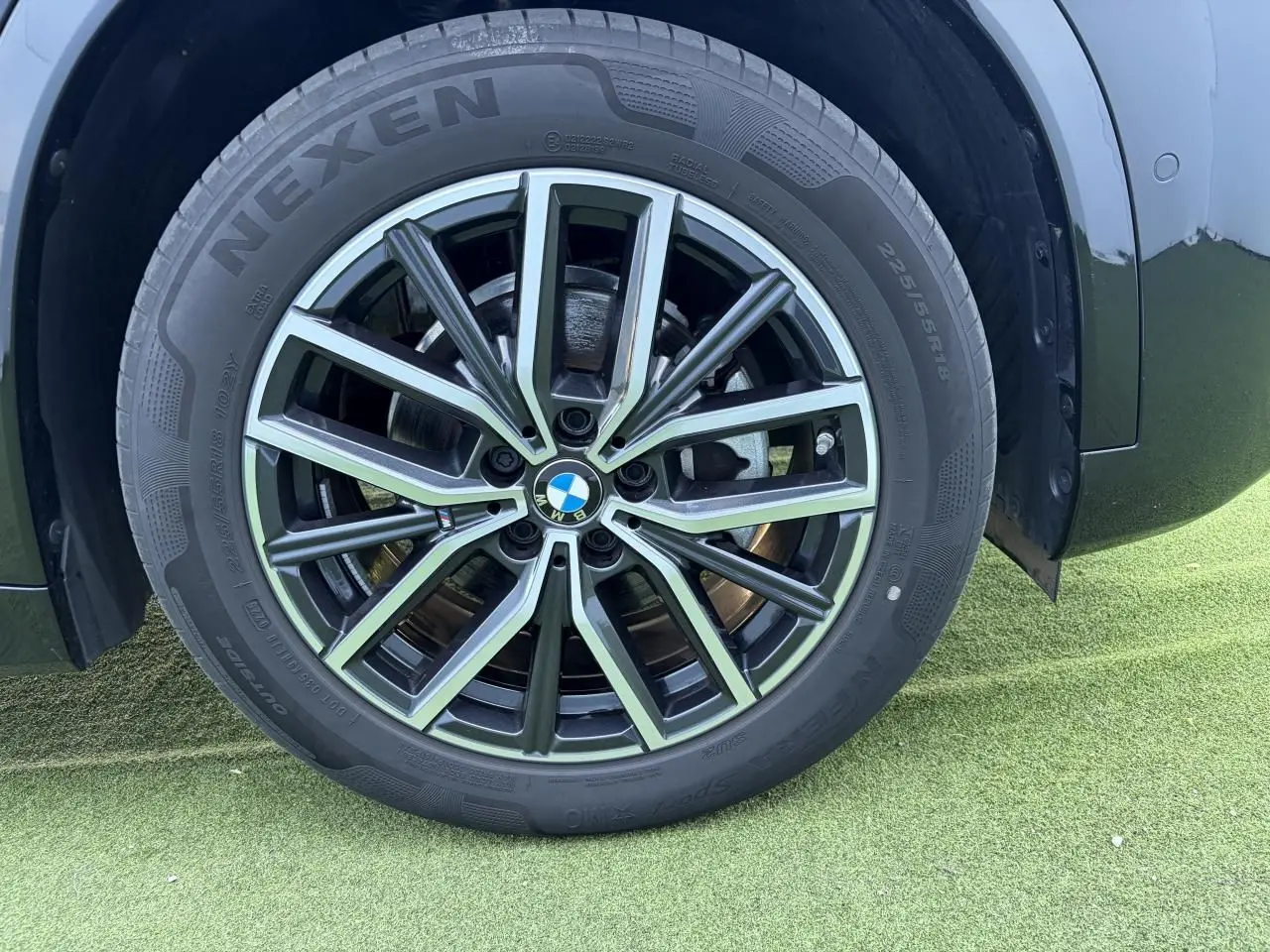 Gros plan sur la jante alliage 18 pouces bicolore du BMW X1 noir saphir, avec pneu Nexen visible sur sol vert.