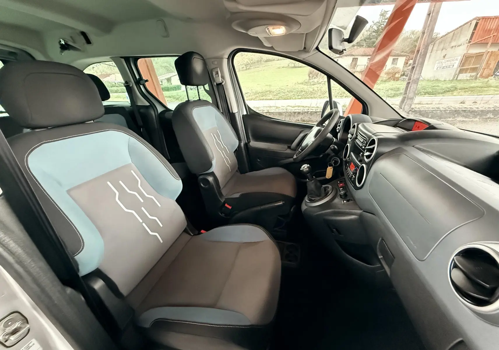 Vue intérieure côté conducteur du Peugeot Partner Tepee 2014, sièges tissu gris et bleu avec volant cuir noir.