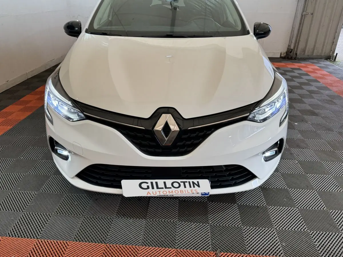 Vue avant d'une Renault Clio blanche 2023 avec phares LED allumés dans un garage au sol damier noir et orange.
