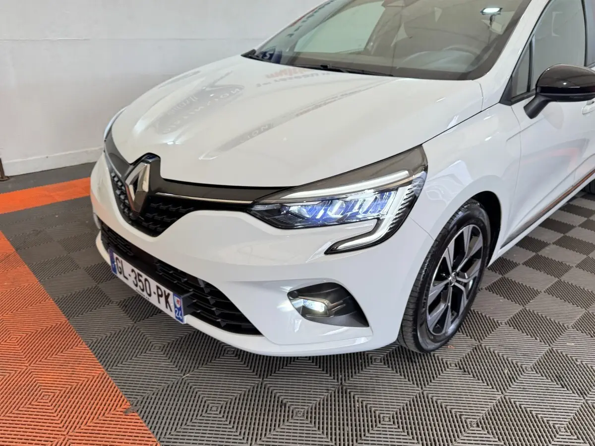 Vue 3/4 avant droite d'une Renault Clio blanche avec phares LED allumés et jantes noires bicolores.