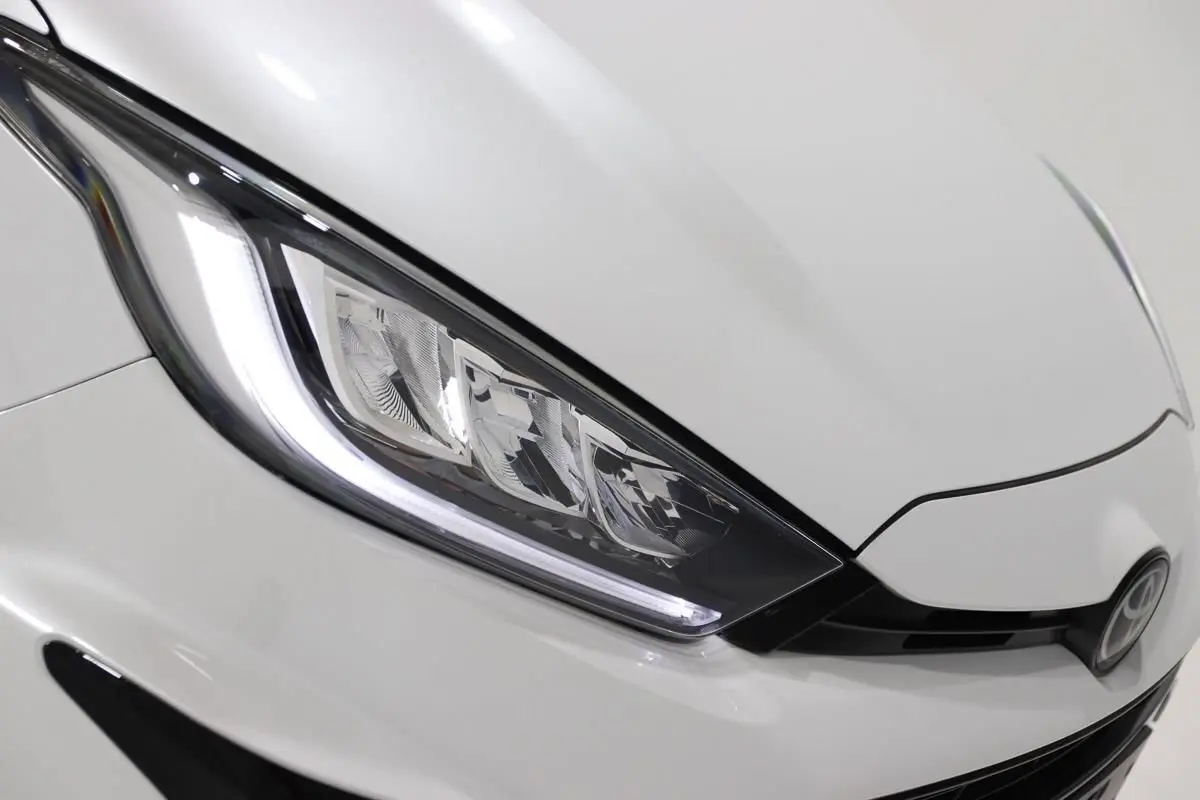 Gros plan sur le phare avant gauche et la calandre noire d'une Toyota Yaris blanc métal.