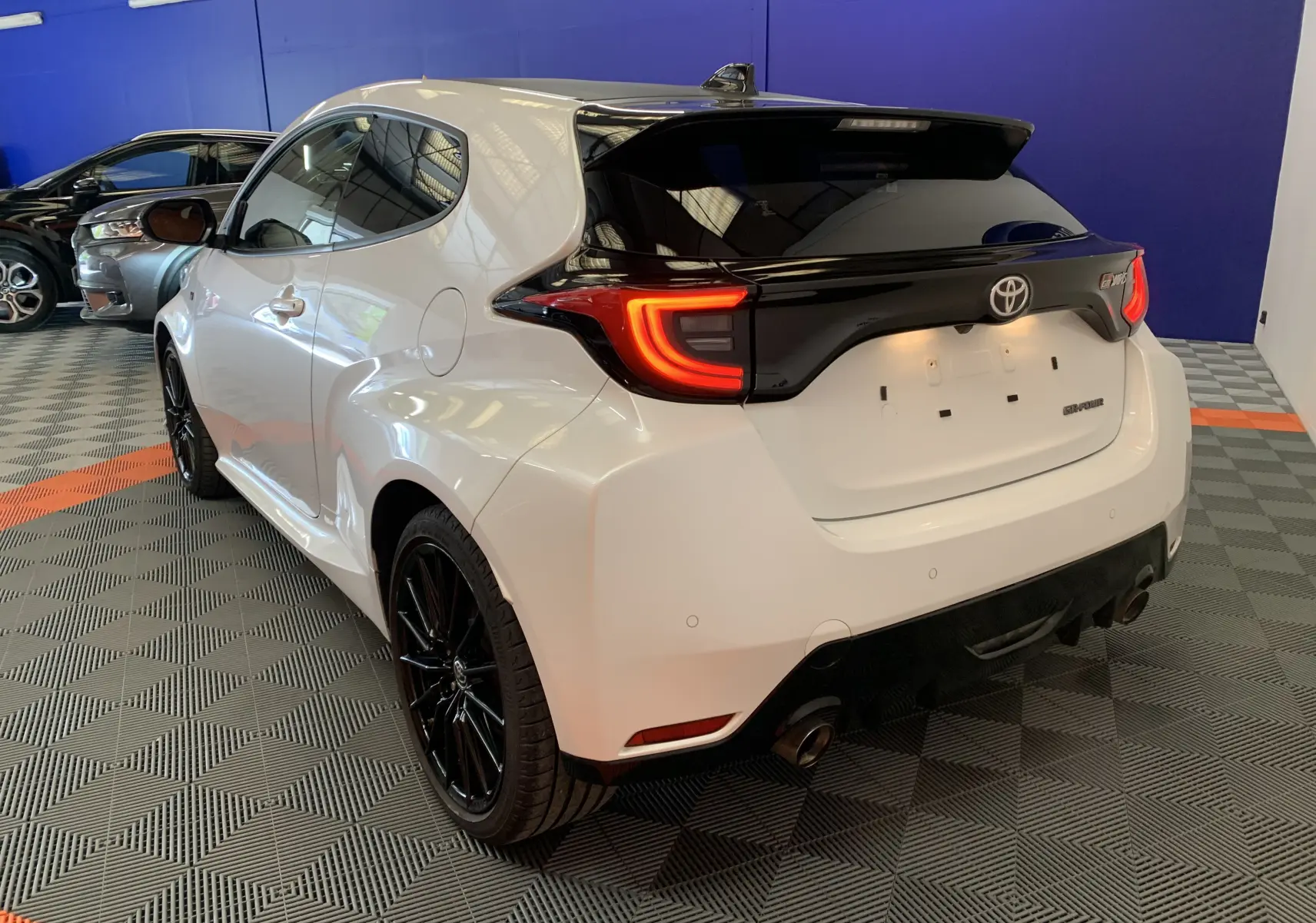 Vue 3/4 arrière droite d'une Toyota Yaris GR blanche métal avec jantes noires et feux arrière allumés dans un showroom.