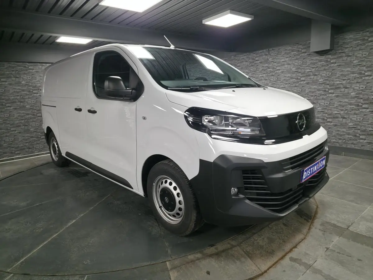 Opel Vivaro blanc en 3/4 avant droit, avec pare-chocs noirs et jantes tôles grises, en intérieur showroom.