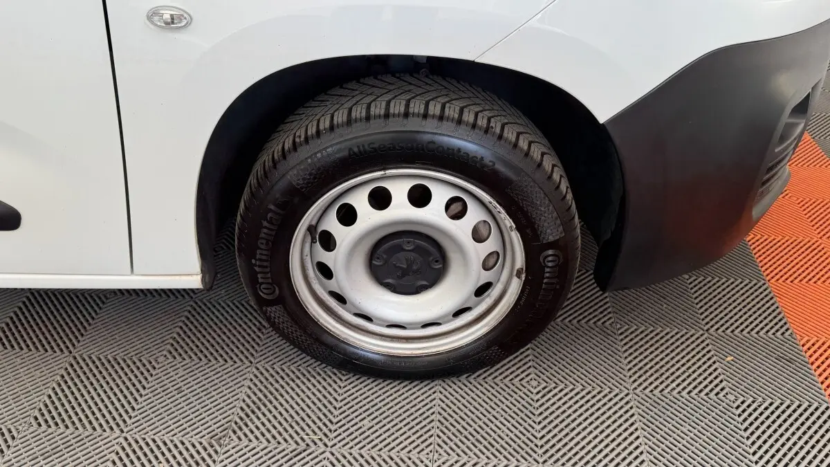 Gros plan sur la roue avant droite acier d’un Peugeot Partner blanc avec pare-chocs noir et sol en damier gris et orange.