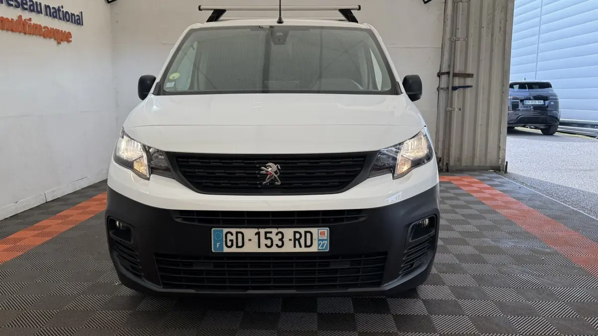 Vue de face du Peugeot Partner blanc avec calandre noire et phares allumés dans un garage.