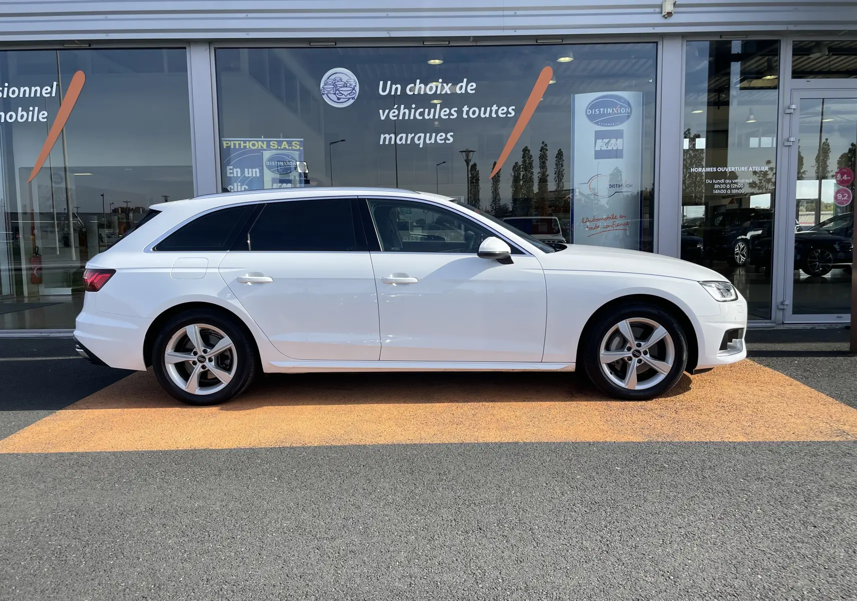 Audi A4 Avant blanc vue de profil côté gauche, garée devant une vitrine de concession multimarque.