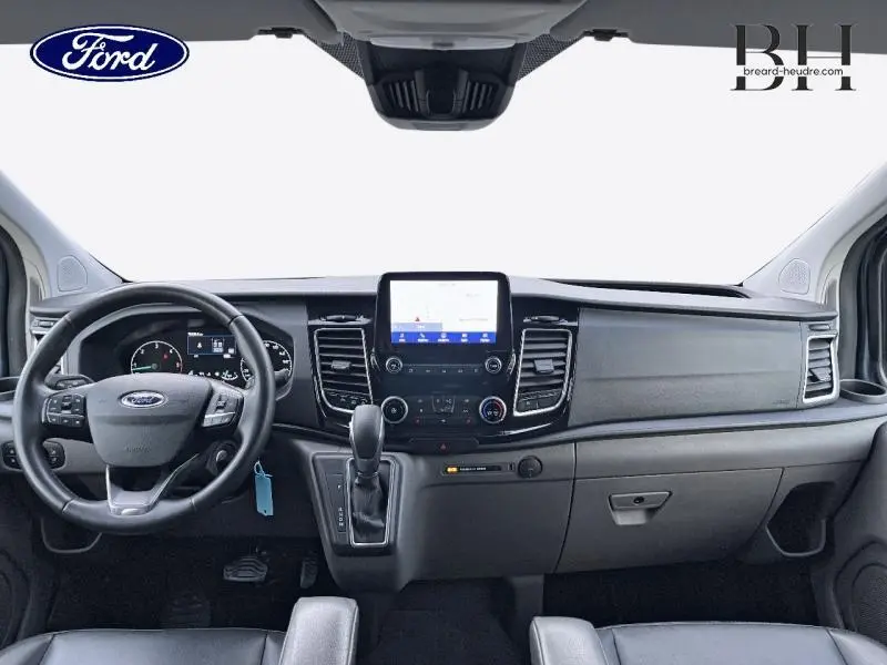 Vue frontale de l'intérieur du Ford Transit Custom Fourgon 2023, tableau de bord noir avec écran tactile central et volant cuir.