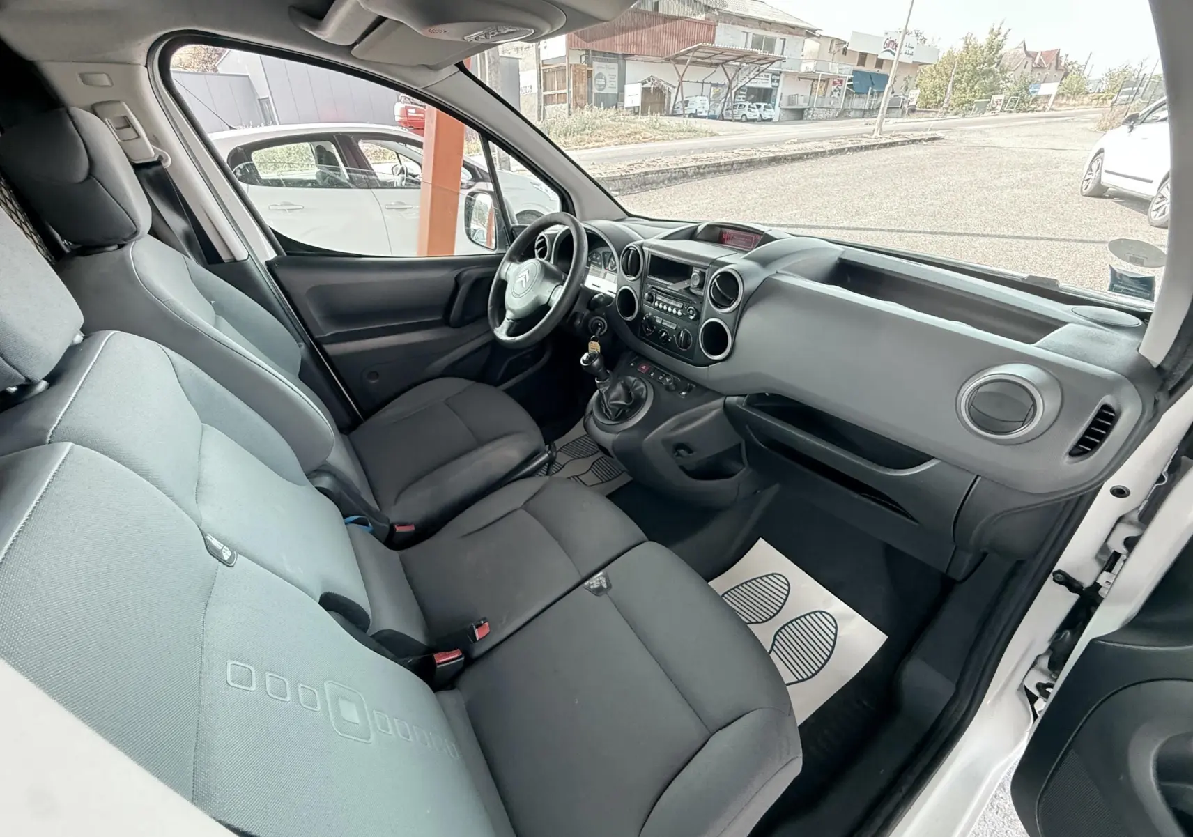 Vue intérieure côté conducteur du Citroën Berlingo blanc, montrant le tableau de bord et les sièges en tissu gris.