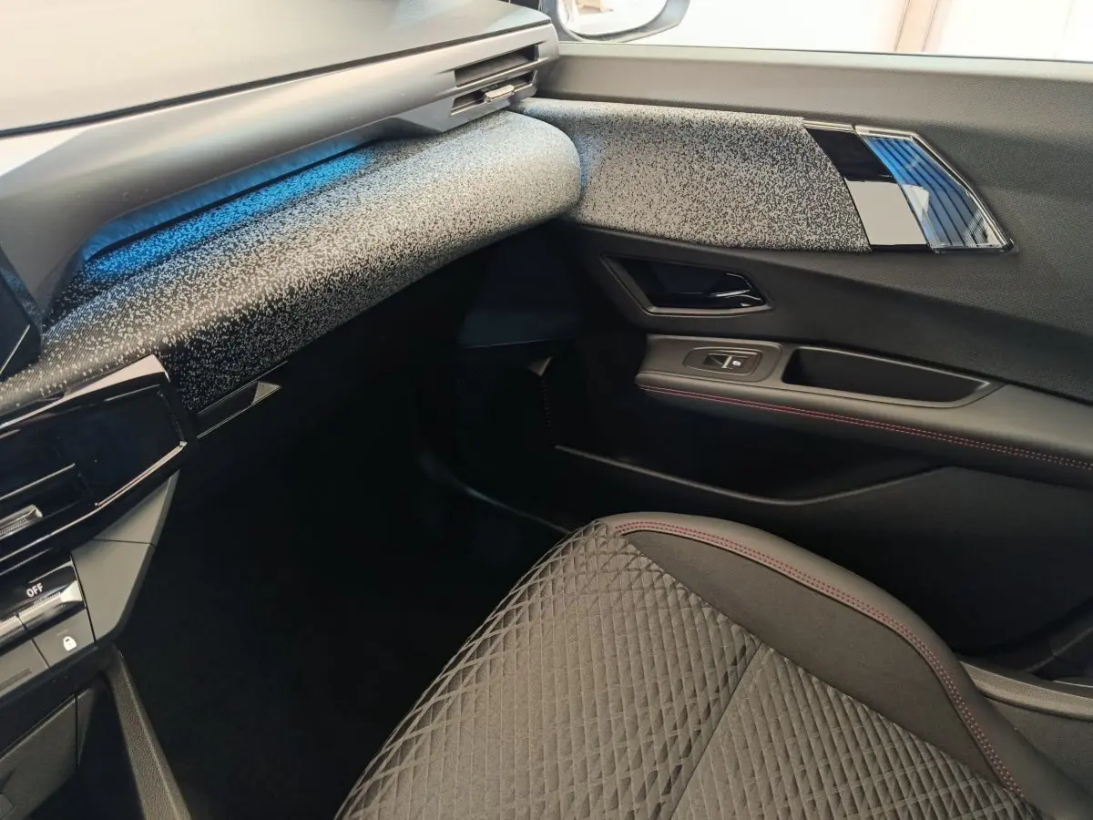 Vue intérieure côté passager avant de la Renault Clio 2026 avec tableau de bord texturé gris et siège tissu noir à surpiqûres rouges.