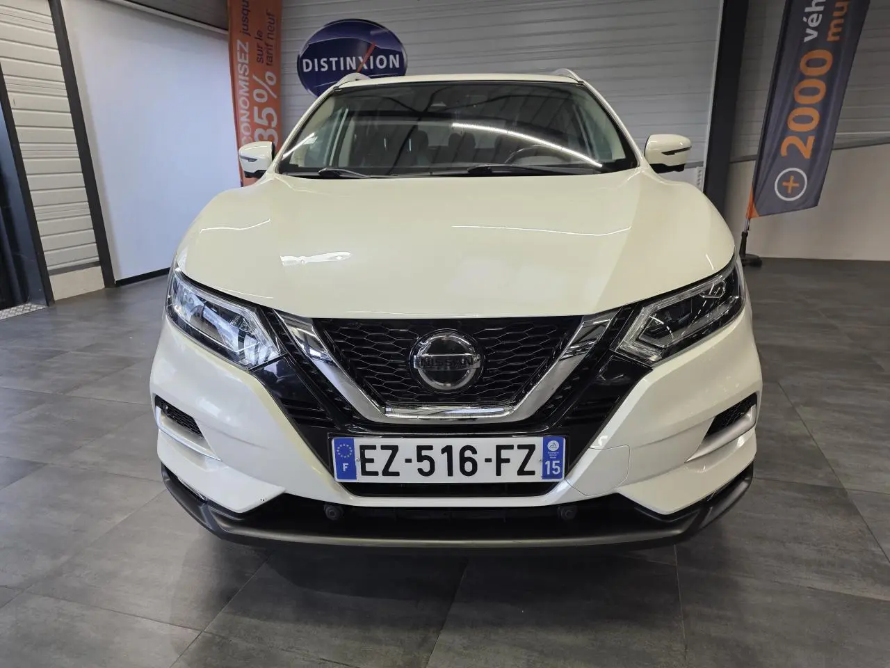 Vue avant d'un Nissan Qashqai blanc avec calandre noire et détails chromés, en intérieur showroom.