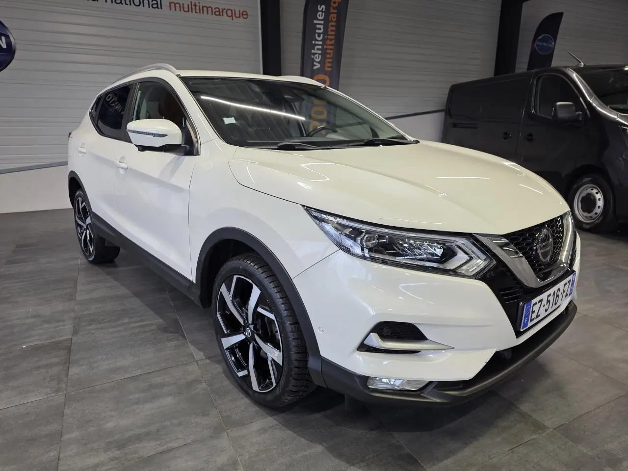 Nissan Qashqai blanc Lunaire en 3/4 avant droit, jantes alliage noires et toit panoramique visible.
