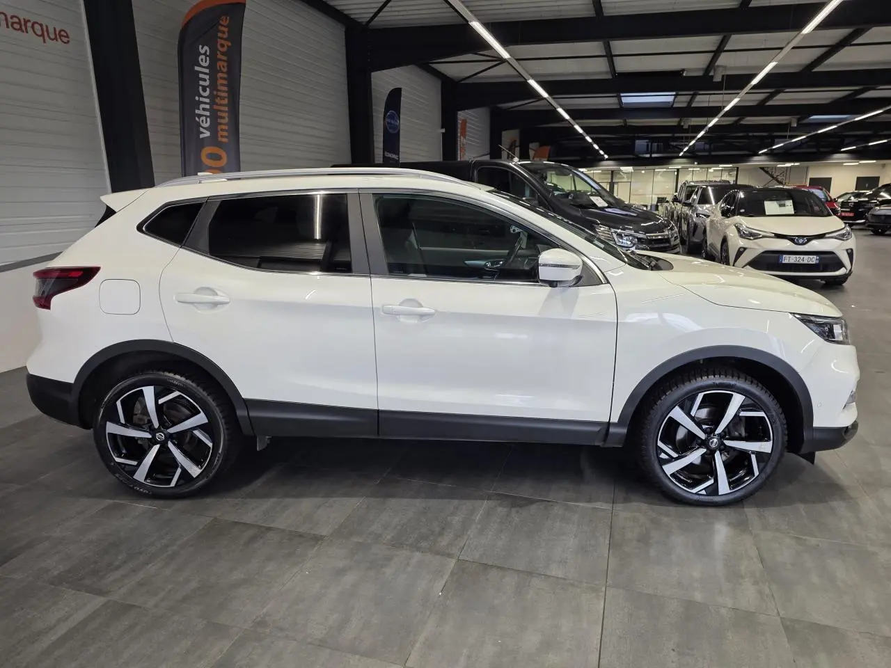 Vue de profil côté gauche d'un Nissan Qashqai blanc avec jantes alliage noires et argent dans un showroom.
