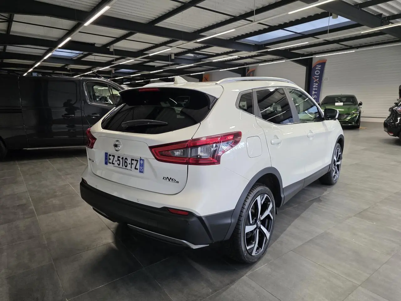 Vue 3/4 arrière droite d'un Nissan Qashqai blanc avec jantes alliage noires et toit panoramique en intérieur showroom.