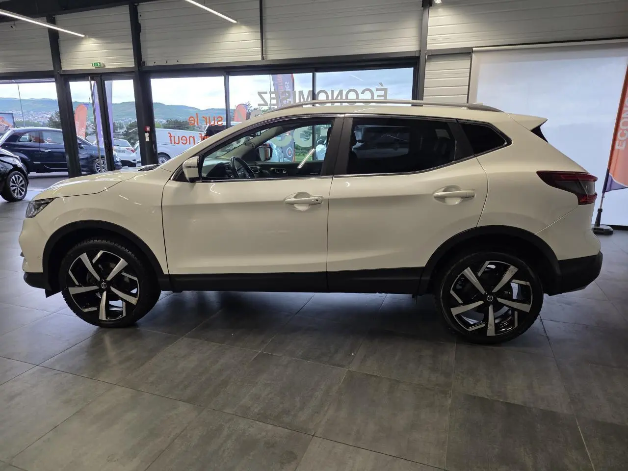 Profil droit du Nissan Qashqai blanc Lunaire avec jantes alliage 19 pouces et toit panoramique en showroom.