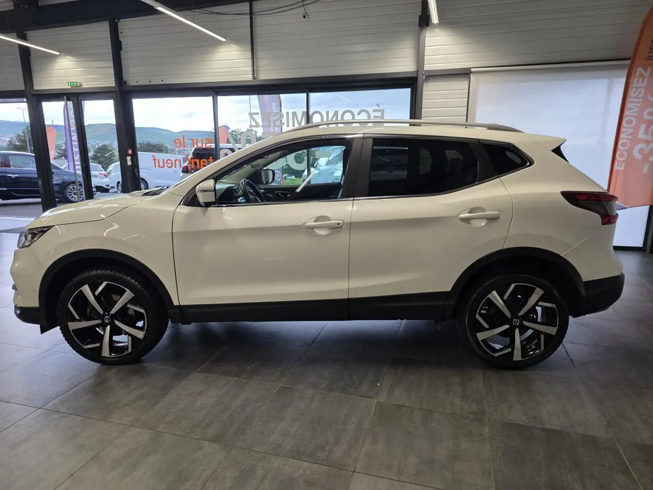 Vue de profil droit du Nissan Qashqai blanc Lunaire avec jantes alliage noires et toit panoramique en showroom.