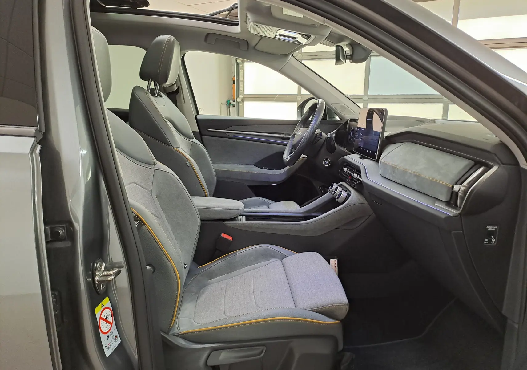 Vue intérieure côté conducteur du Skoda Kodiaq gris graphite 2025, sièges tissu gris avec surpiqûres orange et toit panoramique ouvert.