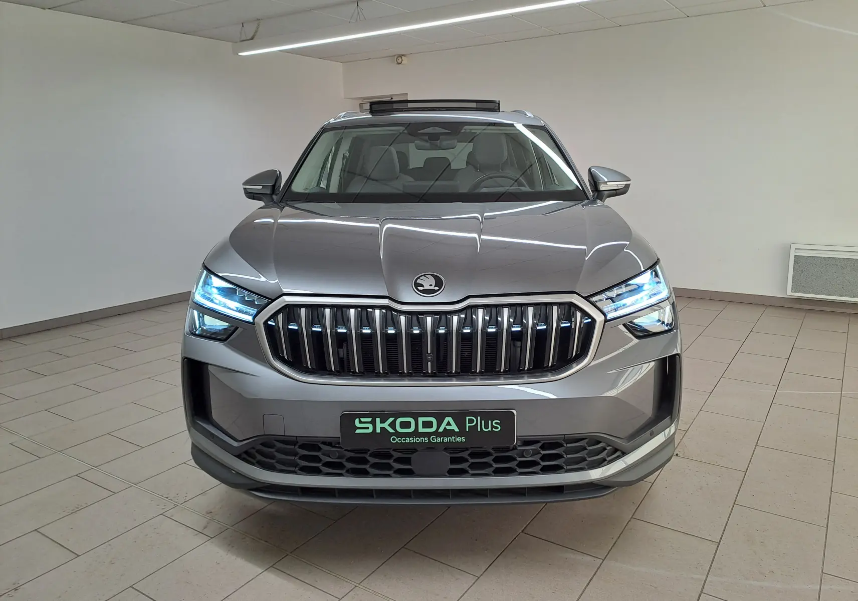 Vue frontale d’un Skoda Kodiaq gris graphite 2025 avec calandre verticale et phares LED allumés dans un showroom.
