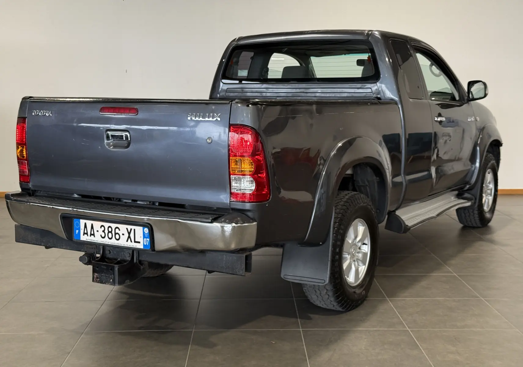 Vue 3/4 arrière droite d'un Toyota Hilux X-Tra Cabine gris 2009 avec benne et pare-chocs arrière légèrement abîmé.
