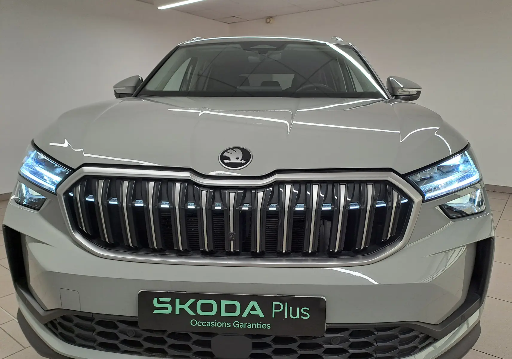 Vue frontale d'un Skoda Kodiaq gris perle avec calandre verticale et phares LED allumés en intérieur showroom.