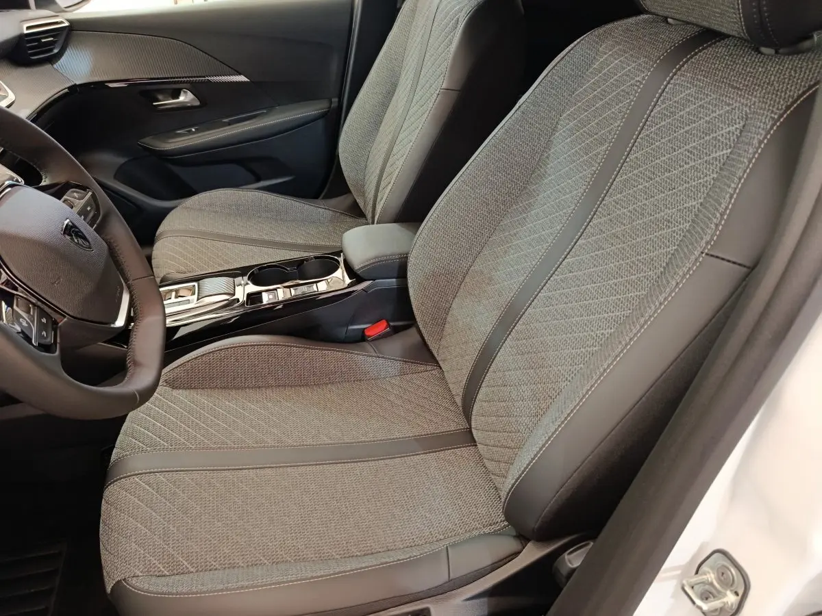 Intérieur de la Peugeot 208 électrique 2024, vue côté conducteur sur sièges tissu gris et console centrale noire brillante.