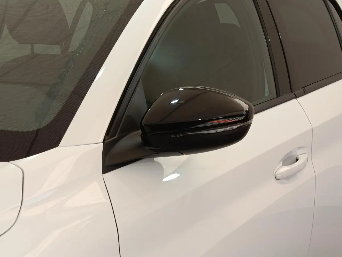 Gros plan sur le rétroviseur noir côté conducteur d’une Peugeot 208 électrique blanche 2024, avec poignée de porte visible.
