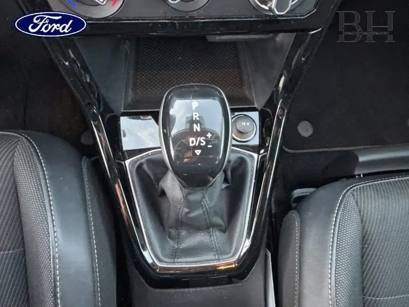 Gros plan sur le levier de vitesses automatique noir brillant entre les sièges avant en tissu gris foncé.