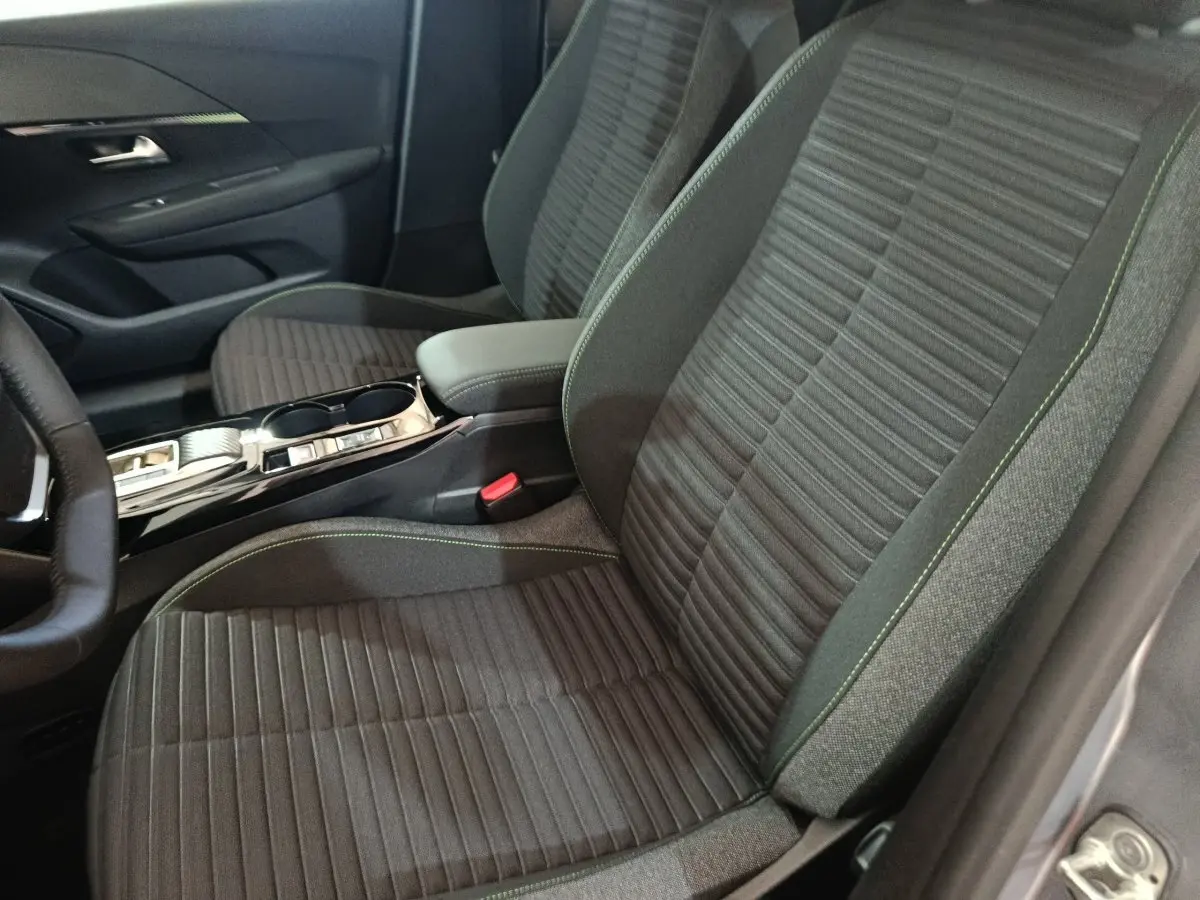 Vue intérieure côté conducteur de la Peugeot 208 électrique 2024, sièges tissu gris avec surpiqûres vertes et console centrale.