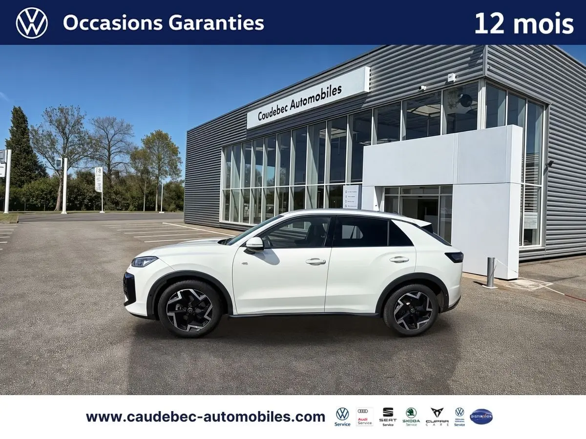 Volkswagen T-Roc blanc vu de profil côté gauche, avec jantes noires et bâtiment de concession en arrière-plan.