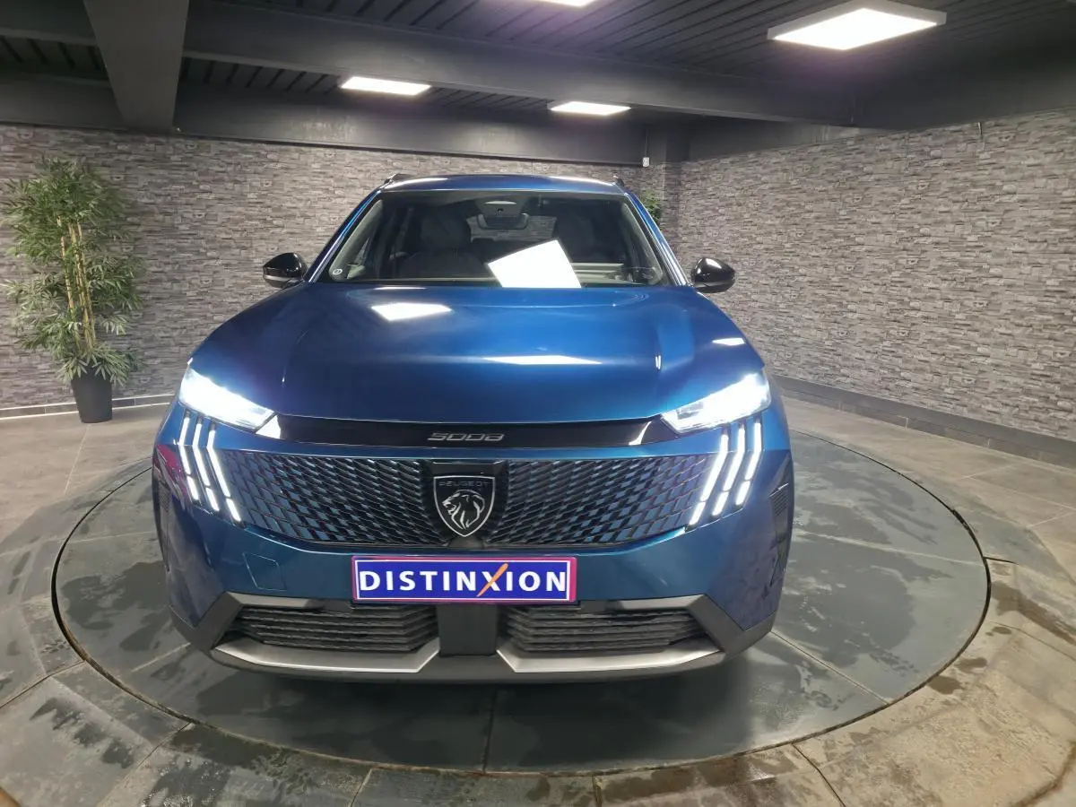 Peugeot 5008 bleu métallisé vue de face, avec calandre noire et feux LED à trois griffes allumés.