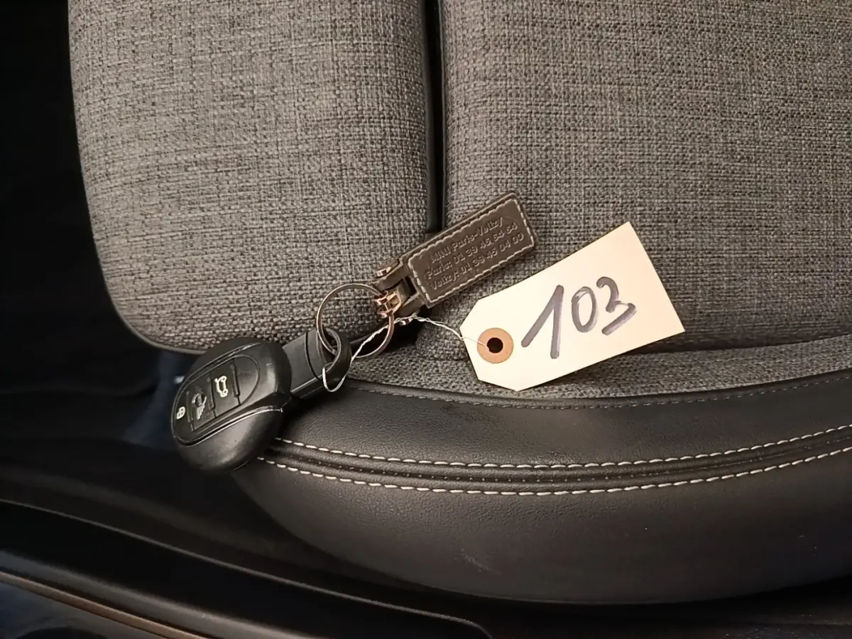 Clé de voiture MINI posée sur un siège tissu gris clair avec bordure cuir noir et étiquette numérotée 103.