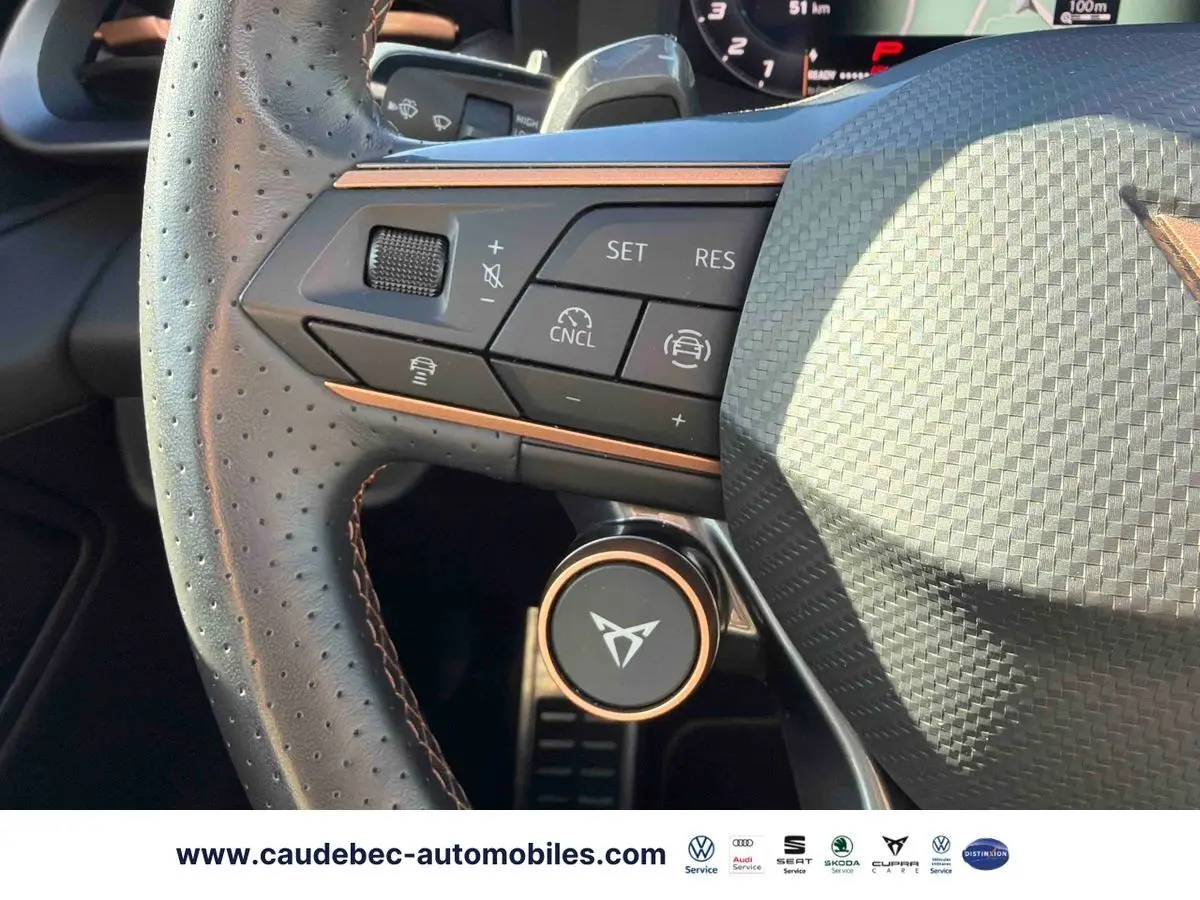 Gros plan sur le volant perforé noir avec commandes et logo CUPRA cuivré, tableau de bord partiellement visible.