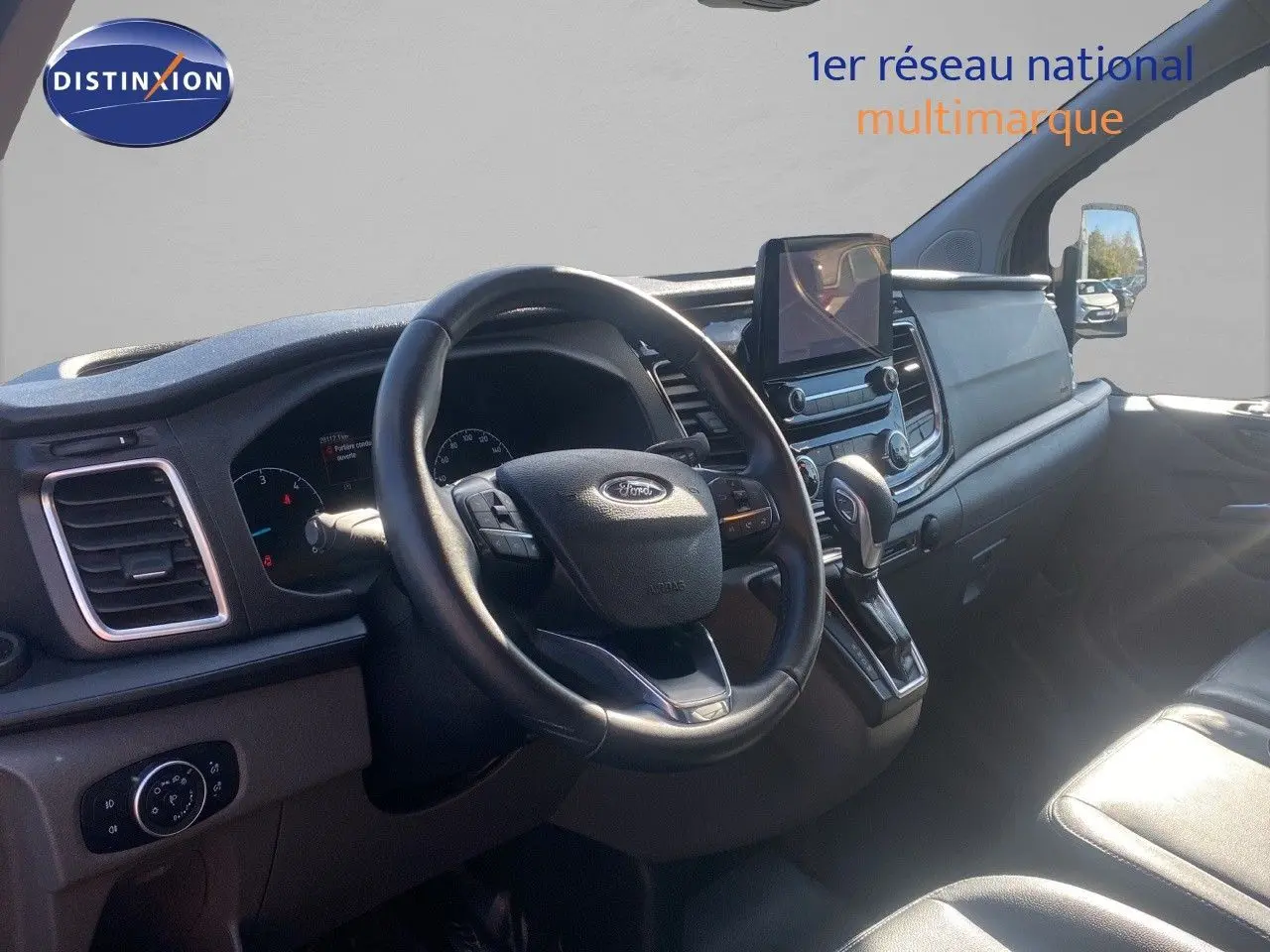 Vue intérieure du poste de conduite du Ford Transit Custom gris, avec volant cuir et écran tactile central.