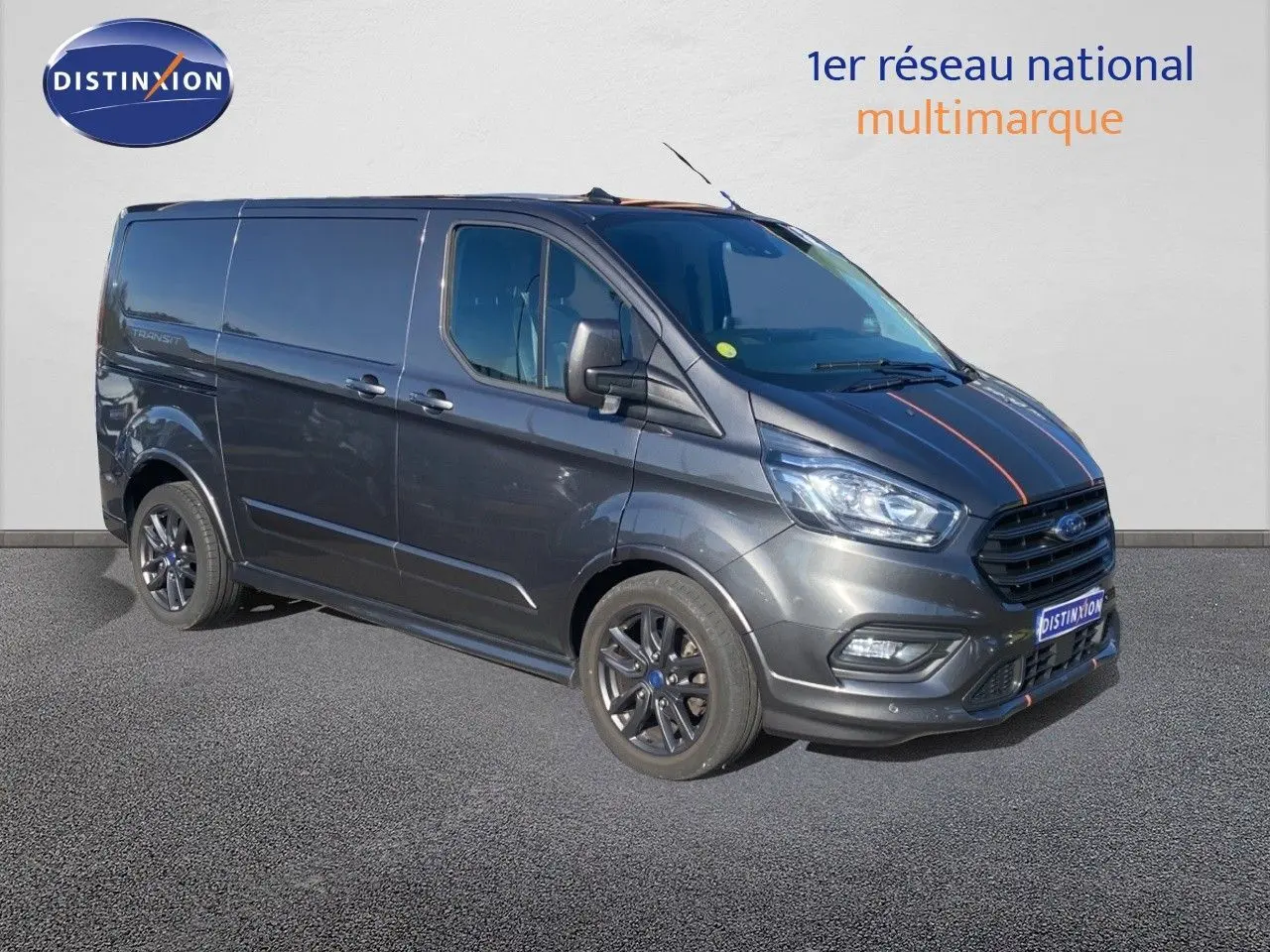Fourgon Ford Transit Custom gris magnetic en 3/4 avant droit avec jantes noires et bandes décoratives rouges sur le capot.