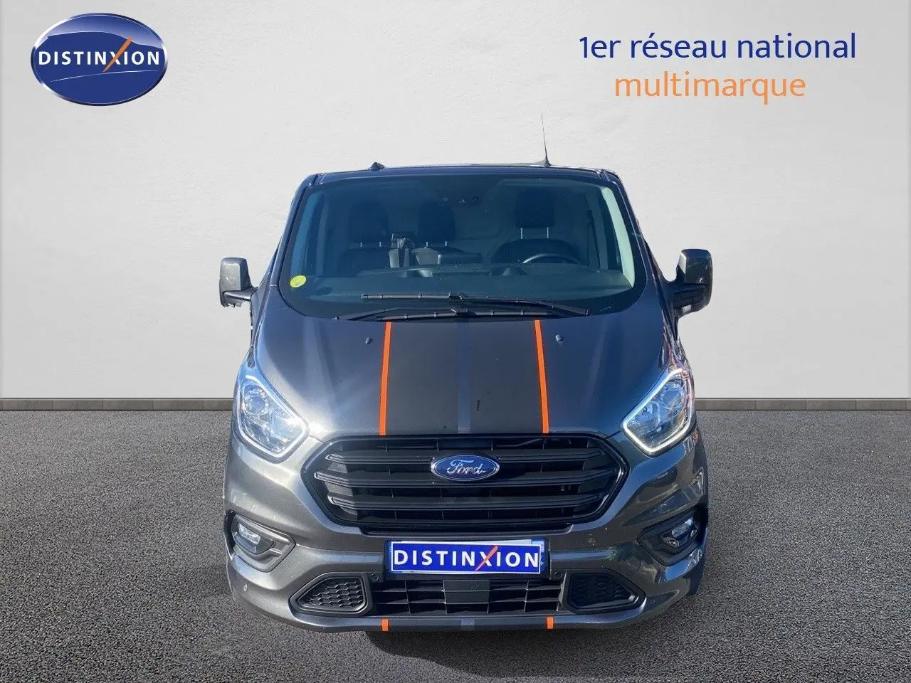 Vue frontale d'un Ford Transit Custom gris magnetic avec bandes noires et orange sur le capot, calandre noire.