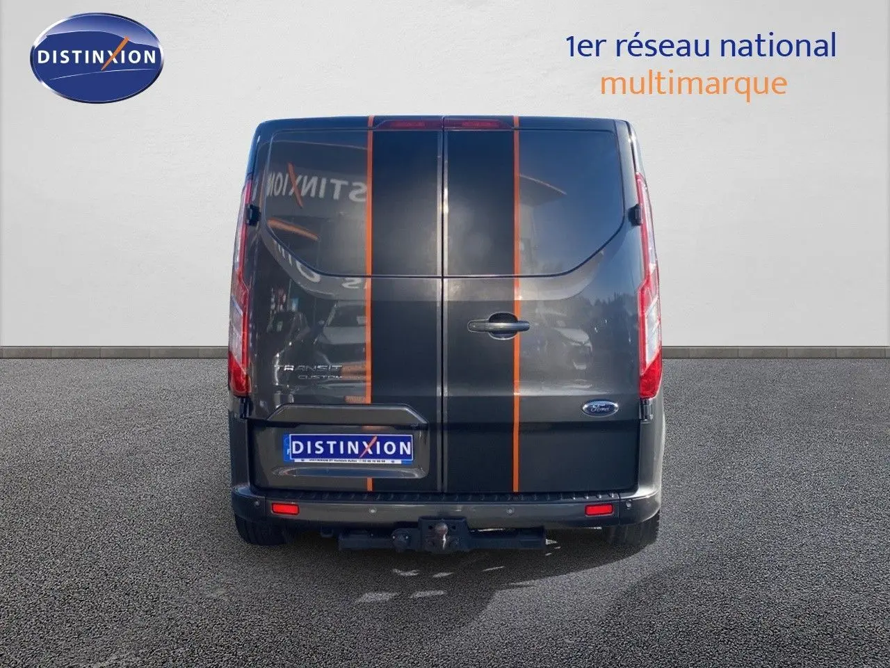 Vue arrière d'un Ford Transit Custom gris magnetic avec bandes orange verticales et attelage visible.