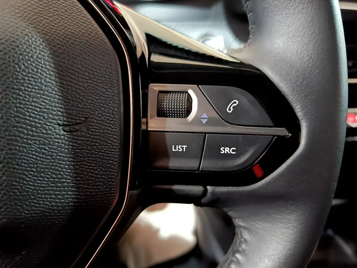 Gros plan sur les commandes gauche du volant cuir de la Peugeot 208 électrique blanche, boutons list, SRC et appel.