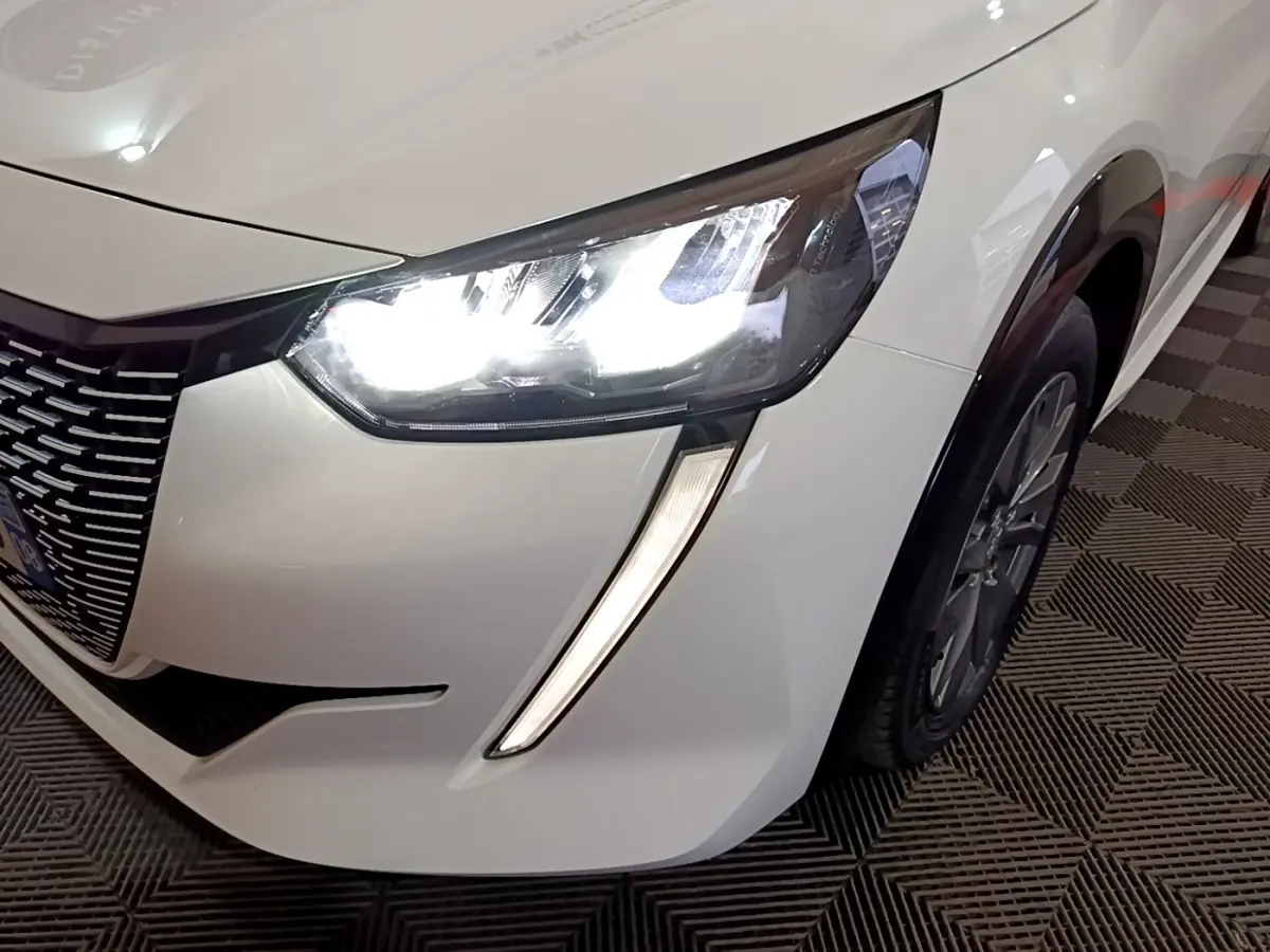 Gros plan sur l'avant droit blanc d'une Peugeot 208 électrique 2021, mettant en valeur le phare LED allumé et la calandre.