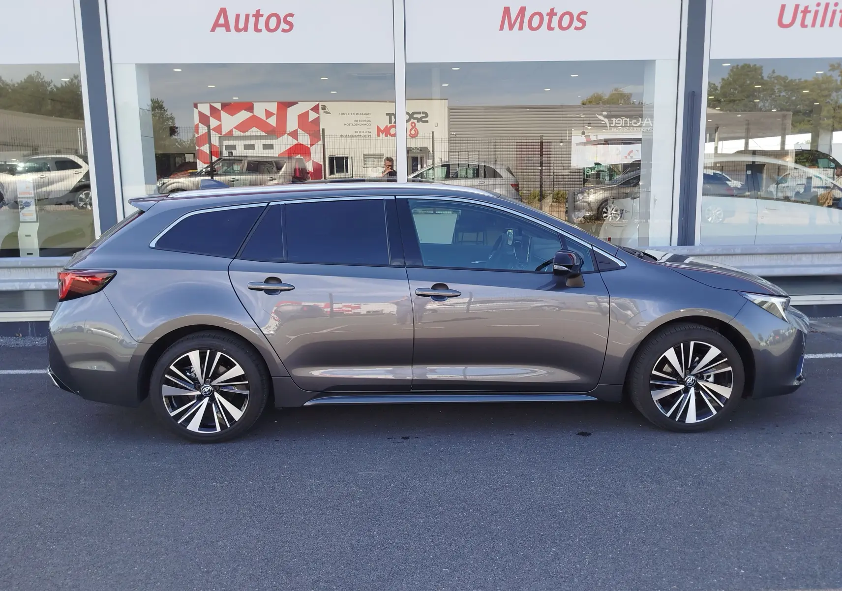 Profil latéral droit d'une Toyota Corolla Touring Sport gris Atlas avec jantes alliage bicolores, stationnée devant une vitrine.