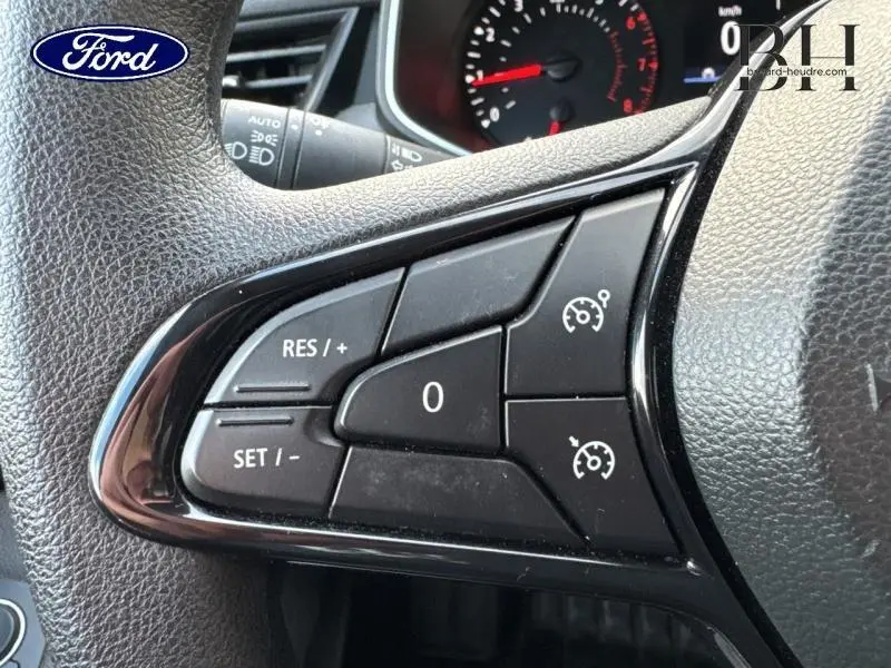 Gros plan sur les commandes du régulateur de vitesse au volant noir d'une Renault Clio blanche, tableau de bord en arrière-plan.